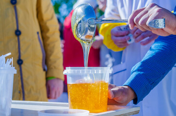 Fair of agricultural products. Woman's cropped hands pour orange bee honey into plastic container.