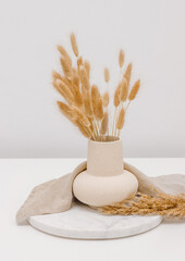 Beige ceramic vase with dried lagurus grass on marble stand with linen cloth, home interior...