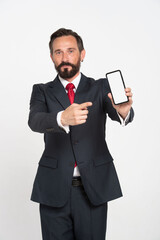 Handsome mature businessman in black suit pointing at smartphone mock up white screen looking at camera isolated on white background. Beard Businessman in suit. App Product placement.