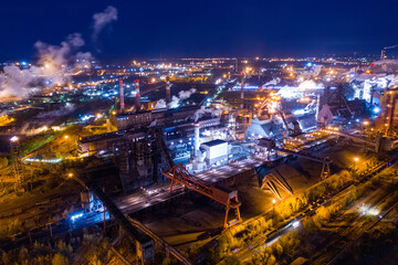 Naklejka premium Aerial view of steel plant at night with smokestacks and fire blazing out of the pipe. Industrial panoramic landmark with blast furnance of metallurgical production. Concept of environmental pollution