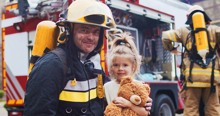 Firefighter hug rescued little girl with teddy bear. Frightened child rejoices in rescue. At background firefighters after extinguishing fire next to fire truck. Concept of saving lives, fire safety