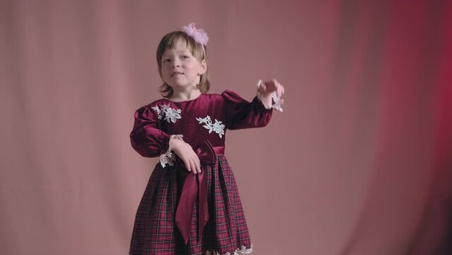 Cute little girl in an vintage burgundy dress is dancing, rejoicing and making funny faces. The concept of children theater productions and developing acting circles. Pink background