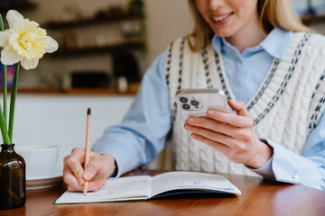 Blonde white woman using mobile phone while writing down notes at cafe