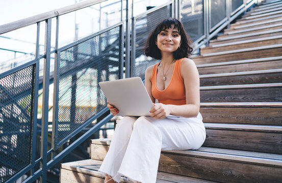 Smiling ethnic woman sitting on steps with laptop and looking at camera