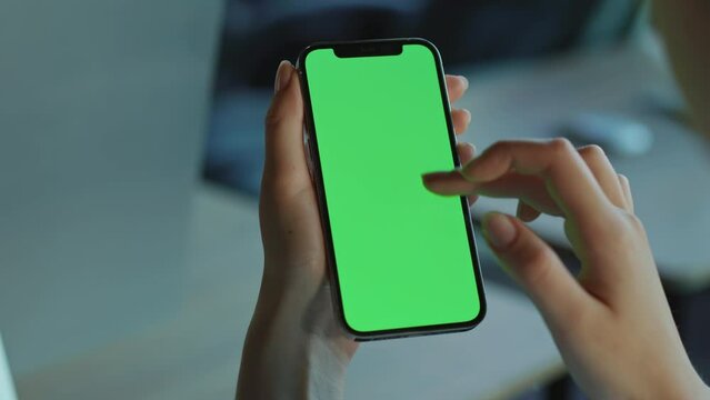 Woman using vertical smartphone with green screen. Female hands holding modern mobile phone in left hand, surfing on Internet on blurred background. Indoors. Gadget, technology