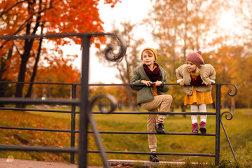 Obraz na płótnie Canvas Autumn park children boy and girl playing on teh bridge in fall golden leaves October season. Kids wearing knitted casual stylish clothes. Positive emotion. Copy space