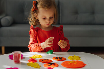 A little girl playing with playdough funny and angry pumpkins and autumn leaves for the holiday of Halloween. Sensory play for toddlers. Holiday Art Activity for Kids.