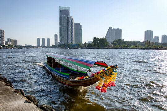 Traditional colorful long-tail boat sailing on the Chao Phraya river surrounded by modern skyscrapers in the background.