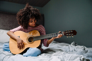 Black girl laughing and playing guitar while sitting on bed
