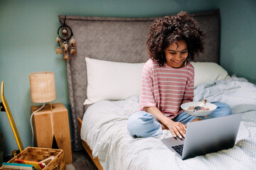 Black girl using laptop and eating cereal while sitting on bed