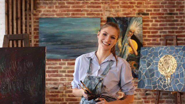 Portrait of young smiling female artist with her artworks in art studio. Painter holding art brushes, hands are stained with paint. Creative process, relaxation, leisure, hobby, stress management.