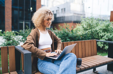 Focused ethnic woman typing on laptop on bench with coffee