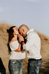 Happy smiling  Couple walking at Sand Dunes near the Beach.  Young happy Bearded muscular  man  in White shirt kissing and hugging beautiful woman at sunser on a beach