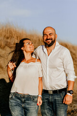 Happy smiling  Couple walking at Sand Dunes near the Beach.  Young happy Bearded muscular  man  in White shirt kissing and hugging beautiful woman at sunser on a beach