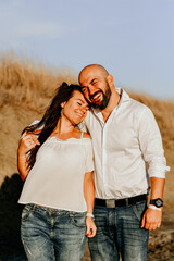 Happy smiling  Couple walking at Sand Dunes near the Beach.  Young happy Bearded muscular  man  in White shirt kissing and hugging beautiful woman at sunser on a beach
