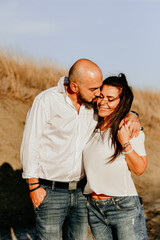 Happy smiling  Couple walking at Sand Dunes near the Beach.  Young happy Bearded muscular  man  in White shirt kissing and hugging beautiful woman at sunser on a beach