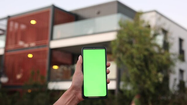Smart home, smart house, application on the mobile phone. Hand holds smartphone with green screen for controlling house security systems.