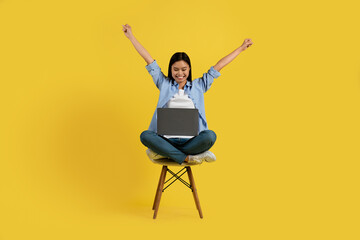 Smiling young chinese female student sits on chair with laptop raises hands up, rejoices to victory