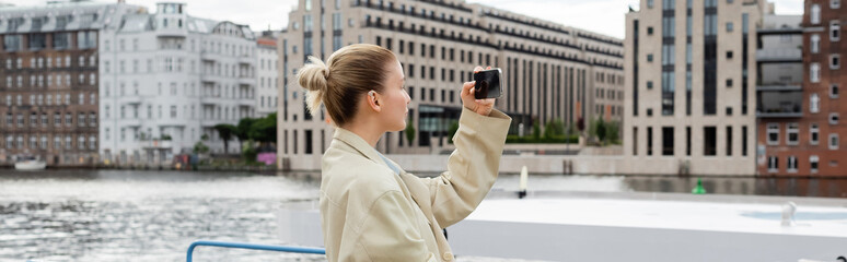 Side view of woman in trench coat taking photo on mobile phone in Berlin, banner.