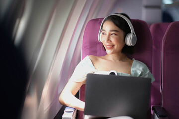 Asian young woman wearing headphone using laptop sitting near windows at first class on airplane during flight, Traveling and Business concept