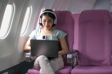 Asian young woman wearing headphone using laptop sitting near windows at first class on airplane during flight, Traveling and Business concept