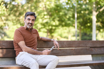 Fototapeta premium Relaxed man looking at the camera and smiling while holding a mobile phone sitting on a park bench. Technology concept.