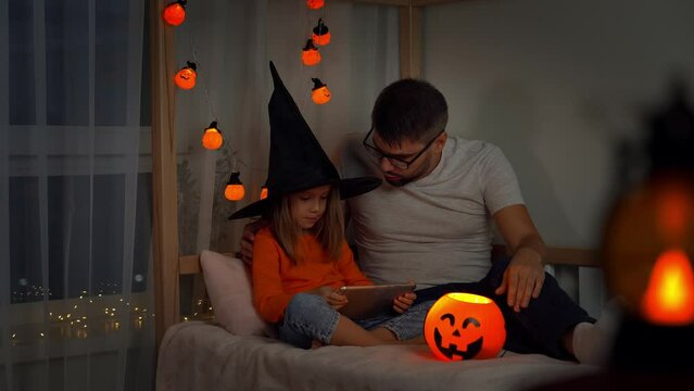 Father and little child girl in witch black hat watch horror film on tablet in dark with orange pumpkin lantern. Halloween