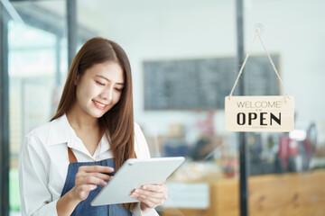 Portrait of a beautiful Asian woman running a small business holding a computer tablet showing a smiling face, a coffee shop owner opening a shop to welcome customers in the new morning, SME concepts