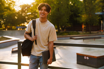 Young handsome smiling asian boy holding backpack and laptop