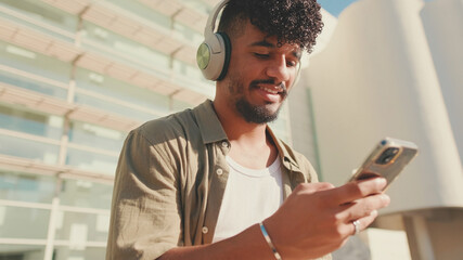 Close up, young male student dressed in an olive color shirt sits outside next to the university, listens to music on headphones, selects tracks on his phone