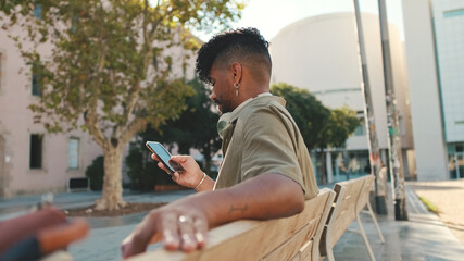 Young man with beard wearing an olive-colored shirt with headphones sits on bench and uses cellphone