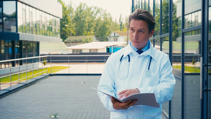 doctor in white coat holding folder with documents and looking away outdoors.