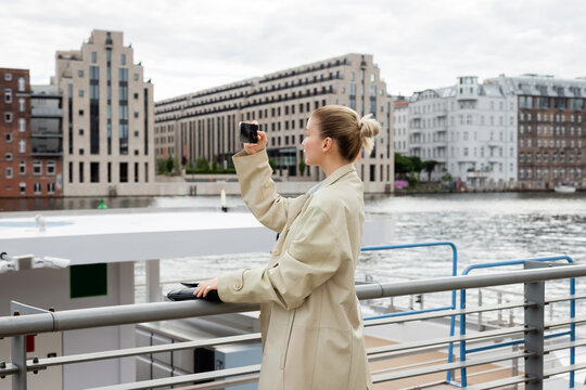 Side view of woman in trench coat taking photo on cellphone near river in Berlin.