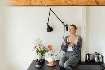 Woman in knitted cardigan holding cup near croissant and flowers in kitchen.