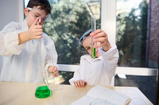 Selective focus on a test tube with green chemical solution in the hands of blurred child girl, watching the going on chemical reaction. Kids learning Chemistry. School laboratory. Knowledge Education