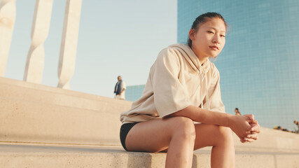 Fototapeta na wymiar Asian girl in sportswear resting after workout while sitting on steps on modern buildings background at sunrise. Young woman turns her head and looks at the camera.