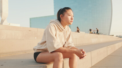 Asian girl in sportswear resting after workout sitting on steps on modern buildings background at sunrise
