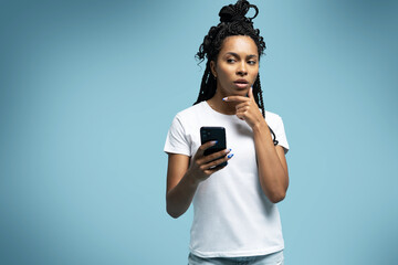 Curly haired ethnic woman uses mobile phone checks messages holds modern cellular in hands isolated...