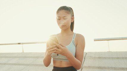 Close-up of smiling asian girl in sports top using cellphone while standing on waterfront on modern buildings background in morning light