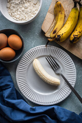 vista superior de un plátano maduro en un plato, un bol con huevos, uno bol con harina y plátanos sobre una tabla de cortar - obrazy, fototapety, plakaty