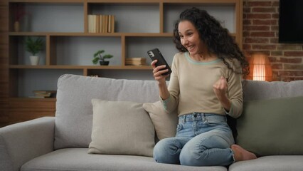 30s Hispanic Latina Caucasian girl woman lady sitting on couch at home living room play with mobile phone win game online betting victory has discount in net virtual hop e-commerce triumph celebrate