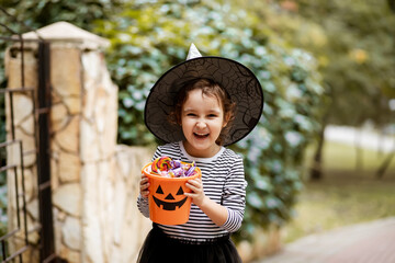 Little cute girl in witch costume holding jack-o-lantern pumpkin bucket with candies and sweets. Kid trick or treating in Halloween holiday. © zulfiska