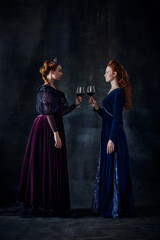 Portrait of two beautiful women in image of queens standing face to face with glass of wine isolated over dark background