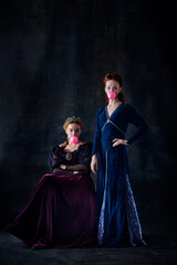 Portrait of two beautiful women in image of queen and princess posing with bubblegum isolated over dark background