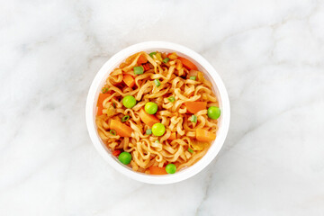 Ramen cup, instant soba noodles in a plastic cup with vegetables, green onions, peas, and carrot, overhead shot on a white marble background