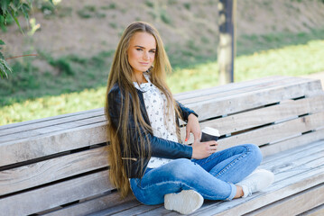 Obraz premium A beautiful fashion young woman sitting on a bench.A girl enjoys coffee from a paper cup.Summer,autumn day.