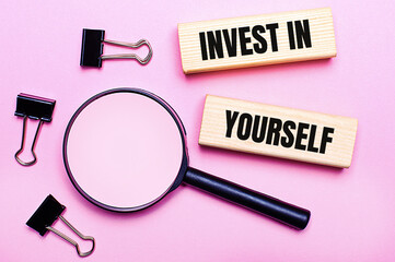 On a pink background, a magnifier, black paper clips and wooden blocks with the text INVEST IN YOURSELF. Business concept