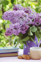 Still life in the spring garden with a bouquet of blooming lilac in a purple pitcher of white polka dot, a stack of books, a Cup of tea and cookies