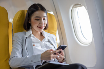Fototapeta premium Traveling and technology. Flying at first class. Pretty young Asian business woman using smartphone while sitting in airplane.