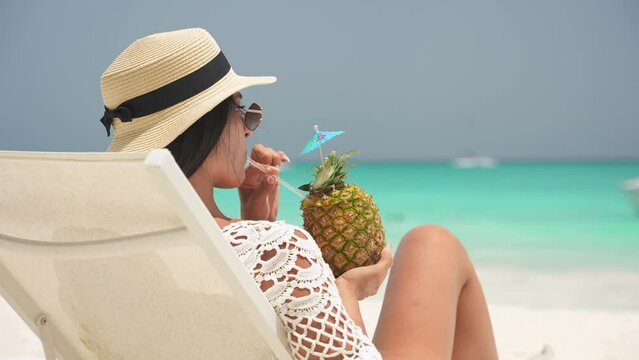Back view of beautiful latin girl on a deck chair at the beach drink pineapple cocktail and enjoys a amazing landscape of the shore of the Caribbean sea. Slow motion shot 